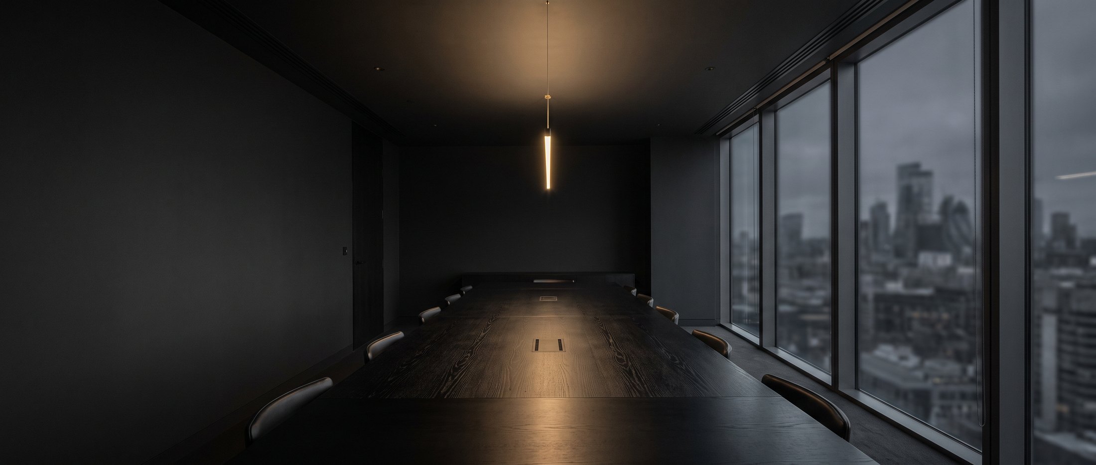 An empty boardroom at night, lit by a single warm pendant, with the London skyline beyond the glass.